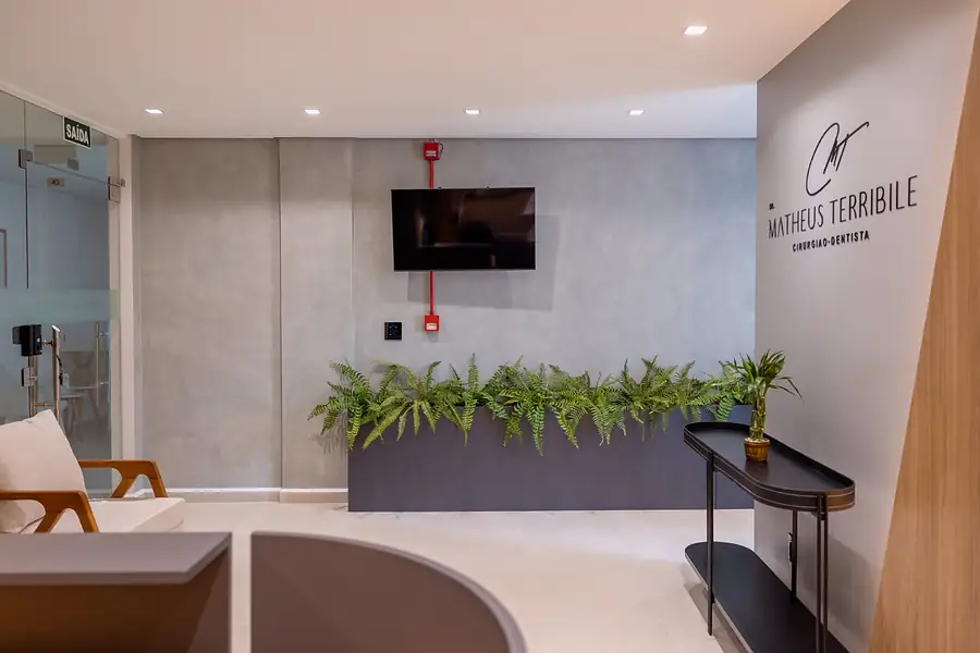 Entrada da clínica, com o nome do Dr. Matheus na parede, jardim de samambaias e sofá claro