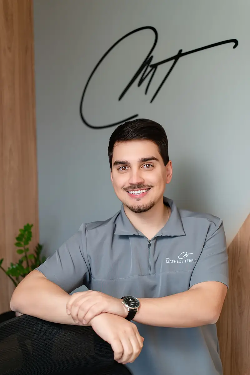 Dr. Matheus Terribile sorrindo, de blusa cinza, com a assinatura da clínica na parede atrás